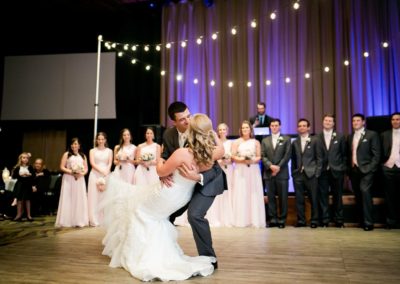 A Groom sweeps his Bride off her feet as they dance and the bridal party looks on at them as they complete their first dance.